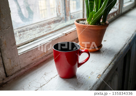 Red mug on an old windowsill next to a houseplant, bringing comfort and warmth to an interior background. Old housing stock 135834336