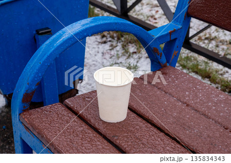 Paper cup on wet wooden bench during light hail or rain. Concept of cold weather, discarded items, urban scene. Paper cup on wet wooden bench during light hail or rain. Concept of cold weather, discarded items, urban scene. 135834343
