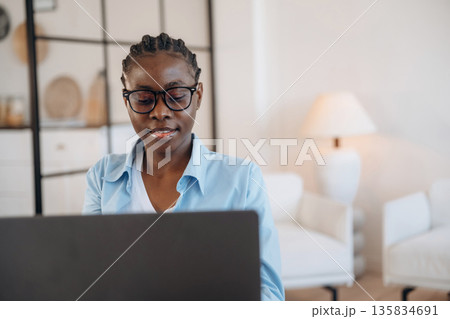 Cheerful, sitting by laptop. Woman is at home in domestic room Cheerful, sitting by laptop. Woman is at home in domestic room 135834691