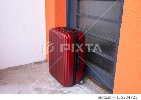 Red rolling suitcase stands next to apartment door. Luggage ready for travel or moving. Departure and arrival concept for journey. 135834725