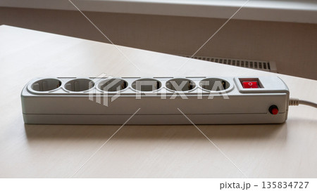 White power extension cable cord with multiple sockets and a red switch on a light wooden table. Electric current distributor for connecting devices. White power extension cable cord with multiple sockets and a red switch on a light wooden table. Electric current distributor for connecting devices. 135834727