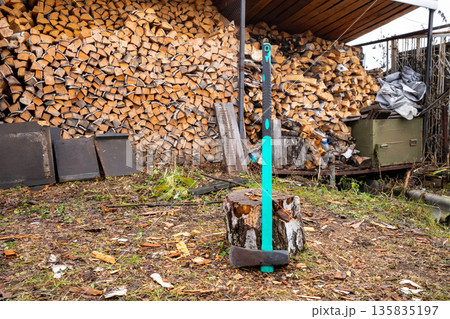 Heavy wood splitting maul stuck in a tree stump with a large pile of stacked firewood in the background. Firewood preparation for winter. Heavy wood splitting maul stuck in a tree stump with a large pile of stacked firewood in the background. Firewood preparation for winter. 135835197
