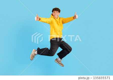 Teen guy jumping mid air with thumbs up gesture on blue background 135835571
