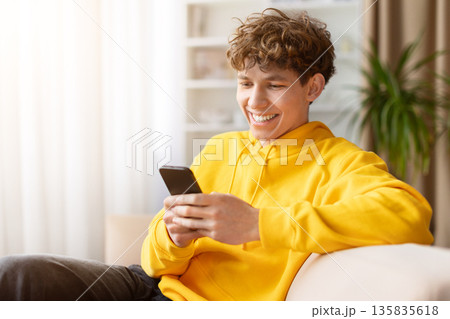 Smiling young man relaxing on sofa while texting on smartphone at home Smiling young man relaxing on sofa while texting on smartphone at home 135835618