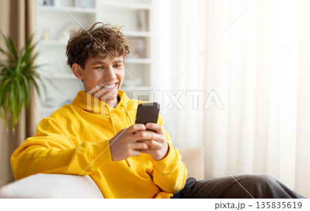 Smiling young man relaxing on sofa and using smartphone at home 135835619