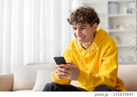 Teen guy at home smiling while texting on smartphone, sitting on sofa 135835645