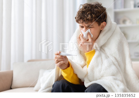 Young man wrapped in blanket at home checking body temperature with thermometer Young man wrapped in blanket at home checking body temperature with thermometer 135835652