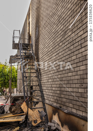 A scorched metal staircase and soot covered brick wall mark the aftermath of a fire, with debris below and caution tape securing the damaged area. 135836620