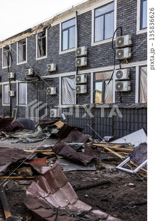 Dark brick building with broken windows and plastic coverings shows signs of recent damage; debris of roof tiles, wood, and metal scattered across the ground in a disrupted urban setting. 135836626