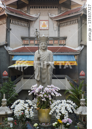Statue of a Buddha figure is surrounded by flowers and plants 135837327
