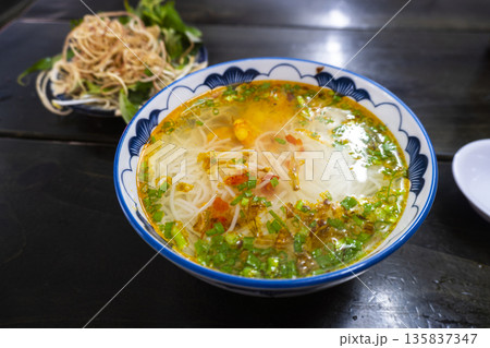 Bowl of soup with noodles and green herbs sits on a table 135837347