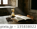 Hourglass measuring time beside old book and beads in historical interior 135838022