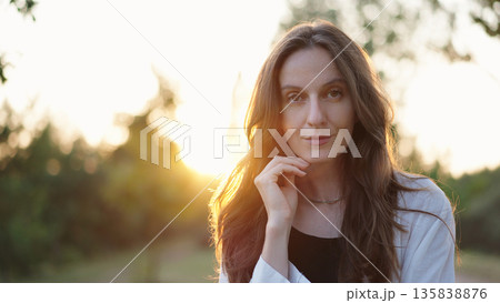 Portrait of a Beautiful Woman Outdoors at Sunset, Female Model Posing Looking at Camera With Hair Swaying in Wind, Gentle Expression, Soft Sun Rays and Nature in Background. Wellness, Mindfulness. Portrait of a Beautiful Woman Outdoors at Sunset, Female Model Posing Looking at Camera With Hair Swaying in Wind, Gentle Expression, Soft Sun Rays and Nature in Background. Wellness, Mindfulness. 135838876