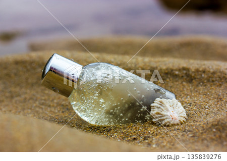 A small, clear, silver-capped cosmetic bottle and a seashell rest on the sand. Bubbles inside the bottle suggest a natural, beach-themed product for skincare. Serum, shampoo, shower gel on a sea coast 135839276