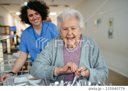 Senior woman having lunch with supportive caregiver in community center cafeteria. Senior woman having lunch with supportive caregiver in community center cafeteria. 135840779