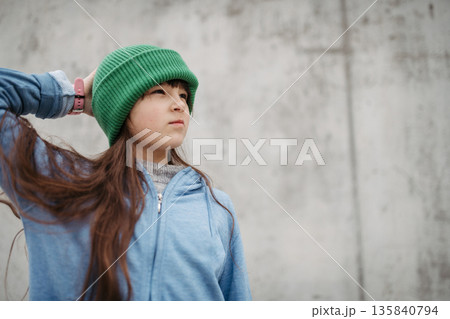 Outdoor portrait of cute young girl with beanie hat. Girl with long hair standing on playground in the city. 135840794