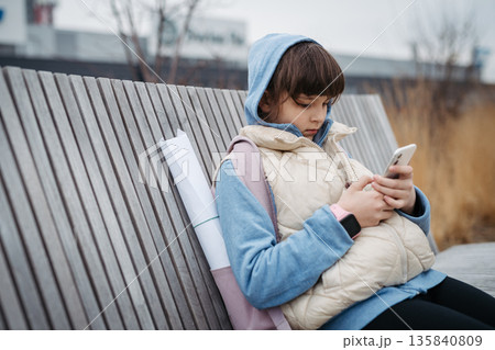 Girl sitting in front of school building, scrolling on phone. Online bullying or cyberbullying for young schoolgirl. Girl sitting in front of school building, scrolling on phone. Online bullying or cyberbullying for young schoolgirl. 135840809