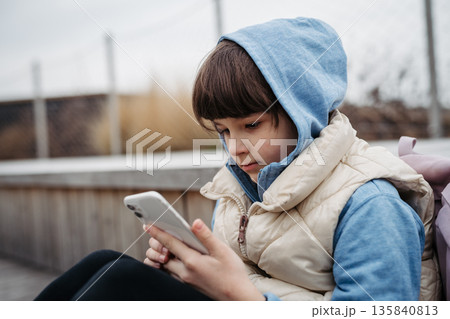 Girl sitting in front of school building, scrolling on phone. Online bullying or cyberbullying for young schoolgirl. 135840813