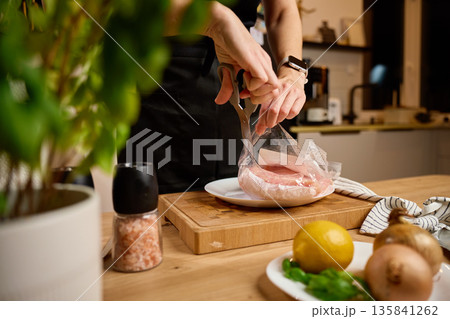 Woman cutting open raw chicken fillet packaging on kitchen counter 135841262