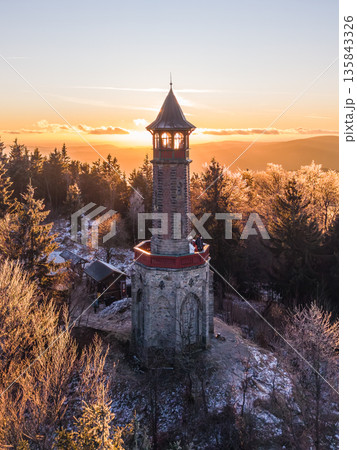 Visitors gather at Stepanka lookout tower in the Jizera Mountains during sunset. The scene features frost-covered trees and a beautiful sky, creating a striking winter atmosphere. 135843326