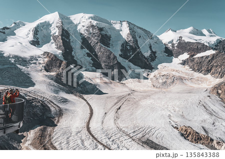 Visitors admire the breathtaking views of Piz Palu and the Pers Glacier while surrounded by towering peaks in the stunning Bernina Range of the Swiss Alps during a clear day. 135843388