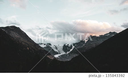 Majestic Piz Bernina rises above the alpine landscape, showcasing a glacier nestled amidst rugged mountains under a dramatic sky at twilight in the Swiss Alps. 135843392
