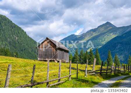 A rustic wooden barn stands beside a winding alpine road in Austria. Surrounded by lush green meadows and majestic mountains, the picturesque landscape captures the essence of rural tranquility. A rustic wooden barn stands beside a winding alpine road in Austria. Surrounded by lush green meadows and majestic mountains, the picturesque landscape captures the essence of rural tranquility. 135843402