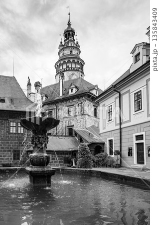 The scene shows a tower rising above buildings in Cesky Krumlov. A fountain is in the foreground with water flowing. It is a summer day in this historic town of Czechia. 135843409
