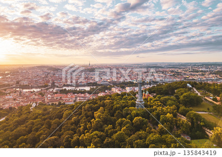 The Petrin Lookout Tower stands majestically at sunrise, surrounded by lush greenery and overlooking the stunning city of Prague, with its historic architecture bathed in golden light. 135843419