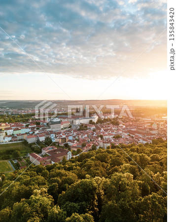 The first light of day casts a warm glow over Prague Castle, illuminating the historic architecture and lush greenery surrounding the city. Morning clouds add depth to the skyline. 135843420
