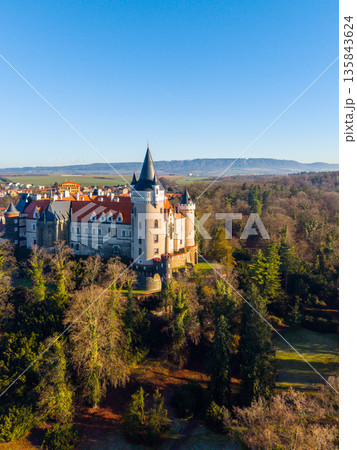 Zleby Castle stands prominently in the landscape, surrounded by trees and open space. The view captures the castle's structure and the surrounding area in bright sunlight. 135843624