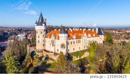 A drone captures Zleby Castle from an aerial perspective. The scene includes surrounding trees and buildings in sunny weather. This location showcases the castle's unique architecture. 135843625