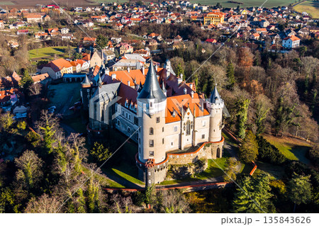 Zleby Castle is shown from the air, surrounded by trees and a small village. The castle has distinctive towers and a red roof. It is located in the countryside. 135843626