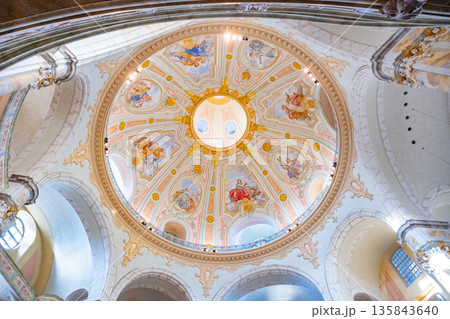 The interior of Frauenkirche in Dresden shows a large dome with detailed paintings. The church features arches and columns. Light enters through windows. Visitors admire the architecture. 135843640