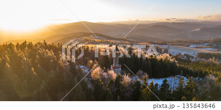 Stepanka lookout tower stands tall in the Jizera Mountains as the sun sets, casting golden light over snowy trees and the landscape below. Visitors enjoy the winter scenery. Stepanka lookout tower stands tall in the Jizera Mountains as the sun sets, casting golden light over snowy trees and the landscape below. Visitors enjoy the winter scenery. 135843646