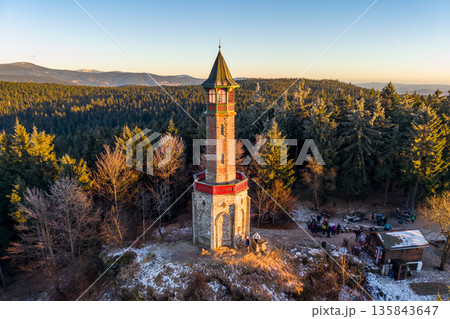 Stepanka lookout tower stands tall in the Jizera Mountains during winter. Visitors gather at the base as the sun sets over the landscape, creating a warm glow in the sky. 135843647