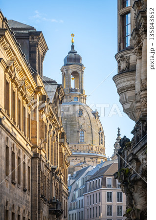 The dome of Frauenkirche rises above the buildings in a narrow street in Dresden. The scene captures the architectural details and historic charm of the area during the day. 135843652