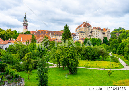 Cesky Krumlov Castle rises above green trees in summer. The castle features distinct architecture and sits within a UNESCO World Heritage site. Visitors enjoy the view and explore the area. 135843658