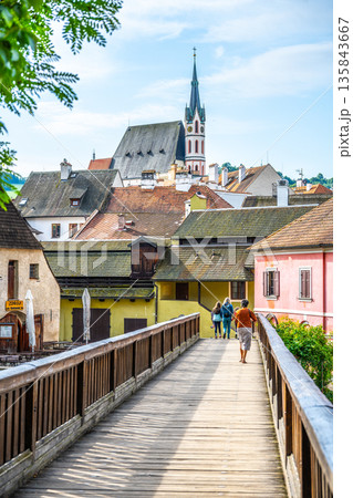 Visitors stroll along a wooden bridge in Cesky Krumlov. The historical buildings and the spire of Saint Vitus Church are visible. The scene captures the town's charm in summer. Visitors stroll along a wooden bridge in Cesky Krumlov. The historical buildings and the spire of Saint Vitus Church are visible. The scene captures the town's charm in summer. 135843667