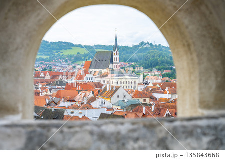 Visitors can see the rooftops of Cesky Krumlov through a stone arch. The town includes buildings with red roofs and a tall church steeple in the background. It is a summer day. Visitors can see the rooftops of Cesky Krumlov through a stone arch. The town includes buildings with red roofs and a tall church steeple in the background. It is a summer day. 135843668