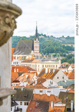 Saint Vitus Church stands tall among red rooftops in Cesky Krumlov. The summer sky brings a soft light to the town's historic buildings and lush greenery in the background. 135843669