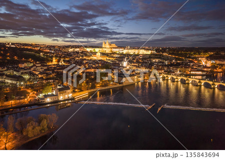 An aerial view of Prague at night. The Vltava River flows beneath the Charles Bridge while the castle is illuminated in the distance. Buildings and streets shine with lights. 135843694