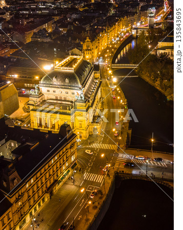 The National Theatre in Prague is illuminated during the evening. The aerial view shows the nearby embankment, river, and city streets filled with lights and activity. The National Theatre in Prague is illuminated during the evening. The aerial view shows the nearby embankment, river, and city streets filled with lights and activity. 135843695