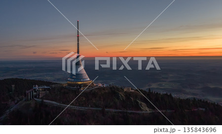 Aerial view captures Jested mountain area at sunset. The hotel stands tall as the sky changes colors. The setting sun creates a beautiful backdrop over the landscape. 135843696
