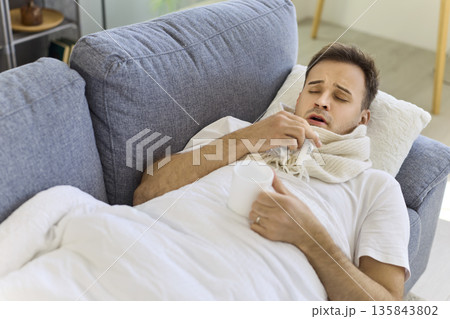 Unwell man with virus lying under blanket on sofa, drinking tea, using tissues Unwell man with virus lying under blanket on sofa, drinking tea, using tissues 135843802