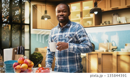Portrait of cheerful young guy holding a cup of fresh coffee at counter, delicious pastry on a plate. Scent of toast and coffee fills the air during morning routine with homemade meal. Portrait of cheerful young guy holding a cup of fresh coffee at counter, delicious pastry on a plate. Scent of toast and coffee fills the air during morning routine with homemade meal. 135844391