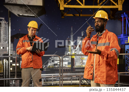 Black drilling rig employee speaking through walkie talkie with colleagues, monitoring oil extraction systems for efficiency. Trying to minimize carbon footprint levels on platform. Black drilling rig employee speaking through walkie talkie with colleagues, monitoring oil extraction systems for efficiency. Trying to minimize carbon footprint levels on platform. 135844437