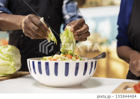 Close up of African american guy ripping and washing lettuce leaves for a salad, adding fresh ingredients and chopped vegetables in a bowl for vegetarian meal. Healthy lifestyle with bio produce. Close up of African american guy ripping and washing lettuce leaves for a salad, adding fresh ingredients and chopped vegetables in a bowl for vegetarian meal. Healthy lifestyle with bio produce. 135844484