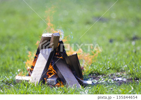 Glowing bonfire on nature. Burning wooden planks outside on summer day. Bright orange flames, light smoke and dark ashes on green grass on blurred green background. Tourism and camping concept. Glowing bonfire on nature. Burning wooden planks outside on summer day. Bright orange flames, light smoke and dark ashes on green grass on blurred green background. Tourism and camping concept. 135844654