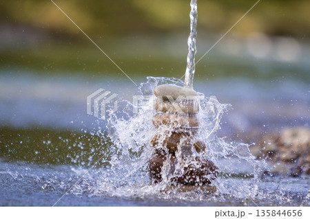 Close-up abstract image of water pouring down on rough natural brown uneven different sizes and forms stones balanced like pyramid pile landmark on blurred blue-green misty copy space background. 135844656